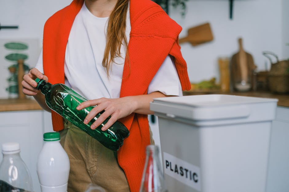A person wearing a white t-shirt and beige trousers is holding a green plastic bottle in their right hand, which is positioned over a white recycling bin labeled 'PLASTIC.' The person has an orange kn