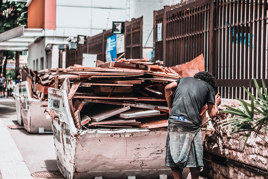A man with curly dark hair, wearing a dark T-shirt and a plaid kilt, stands next to a large, white, metal skip filled with construction debris including wooden planks, tiles, and metal sheets. The deb